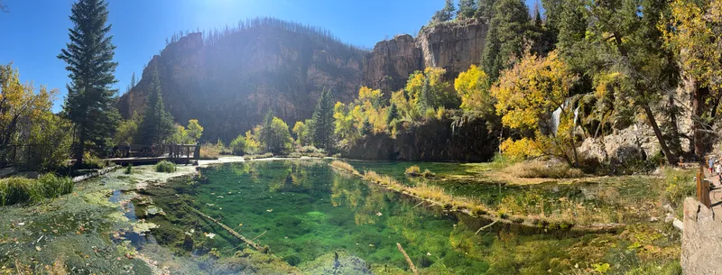 Panorama of Hanging Lake in early October, the turquoise lake ringed by travertine, cattails and a wooden viewing deck beneath tall canyon cliffs.